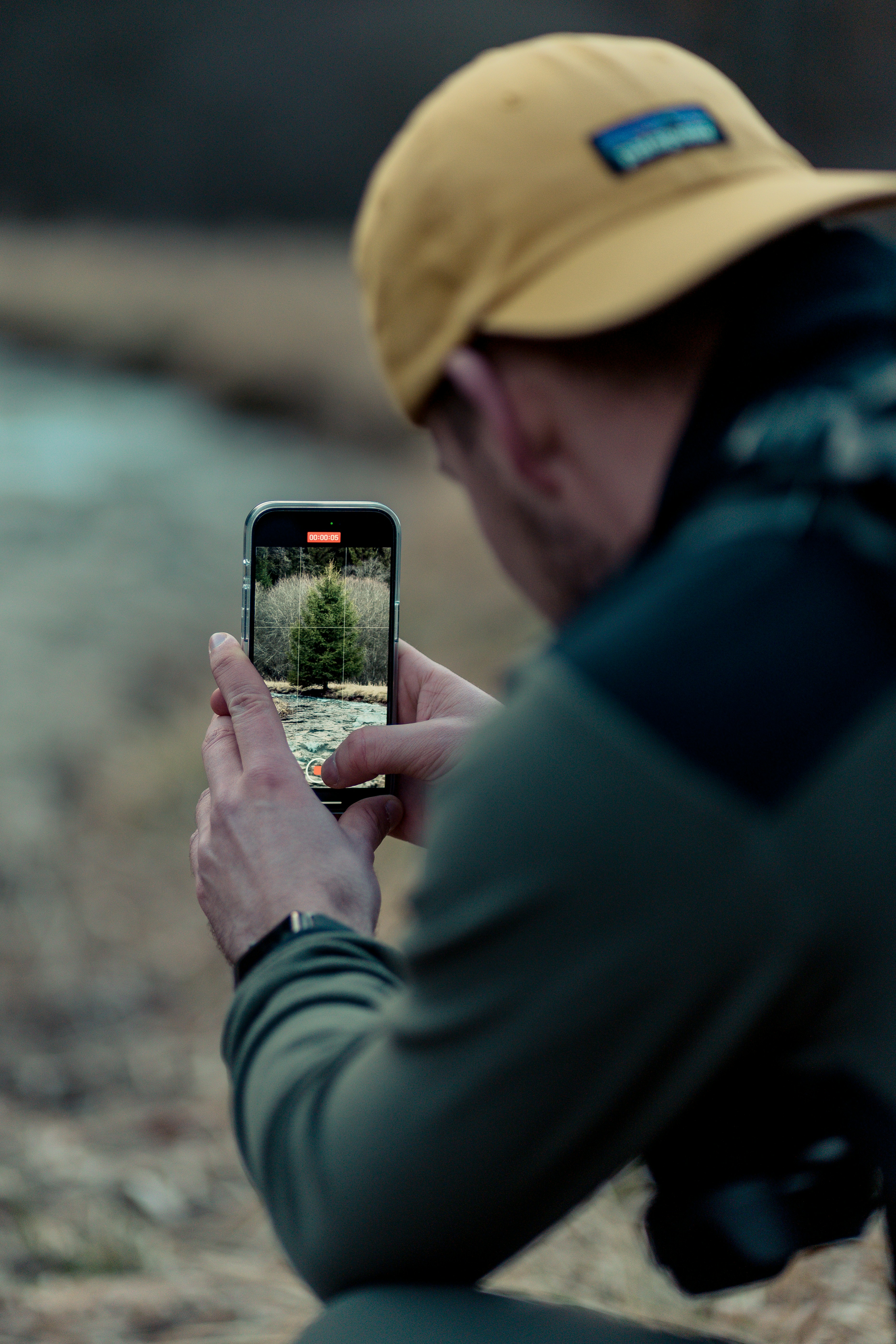 Male with yellow baseball cap taking a picture with his phone of a tree with the  iNaturalist app. 